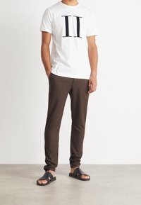 Camiseta blanca de manga corta con un gran estampado negro "II", combinada con pantalones ajustados marrón oscuro y sandalias negras tipo slip-on. Diseño simple y casual.