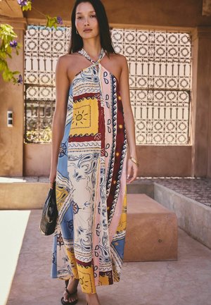 Woman wearing a colorful patchwork halter maxi dress, holding a black handbag, standing outdoors in a sunny, patterned tile courtyard.