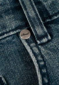 Tela de mezclilla azul oscuro con detalles de costura visibles y un botón de metal con "Reell" grabado. Primer plano de los elementos del diseño de la prenda.