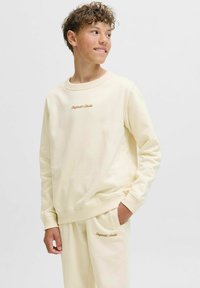 Sweatshirt de cor creme com punhos e decote canelados, apresentando o logótipo bordado "Original Studio" em castanho no peito.
