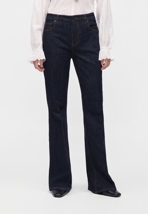 Personne portant un jean large bleu foncé et un chemisier blanc à manches longues avec des poignets volantés, debout sur un fond clair uni.