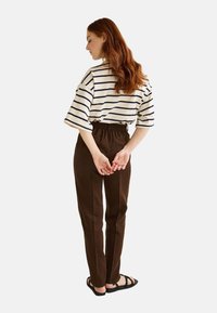 T-shirt en coton à rayures, à manches courtes, de couleur crème et marine, associé à un pantalon brun taille haute, fuselé ; personne de dos, les mains derrière le dos.