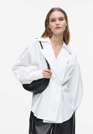 Kobieta w oversize białej marynarce i czarnych skórzanych spodniach, niosąca czarną torebkę na ramię w kształcie półksiężyca ze złotym logo.