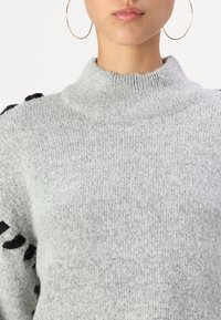 Pull à col roulé gris clair en maille texturée, avec des accents noirs sur les épaules. Tissu lisse avec un col côtelé.