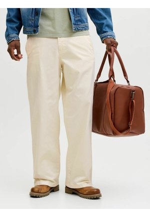 Person iført cremefarvede bukser, brune støvler, denimjakke, bærende en stor brun læder duffel taske i hånden.