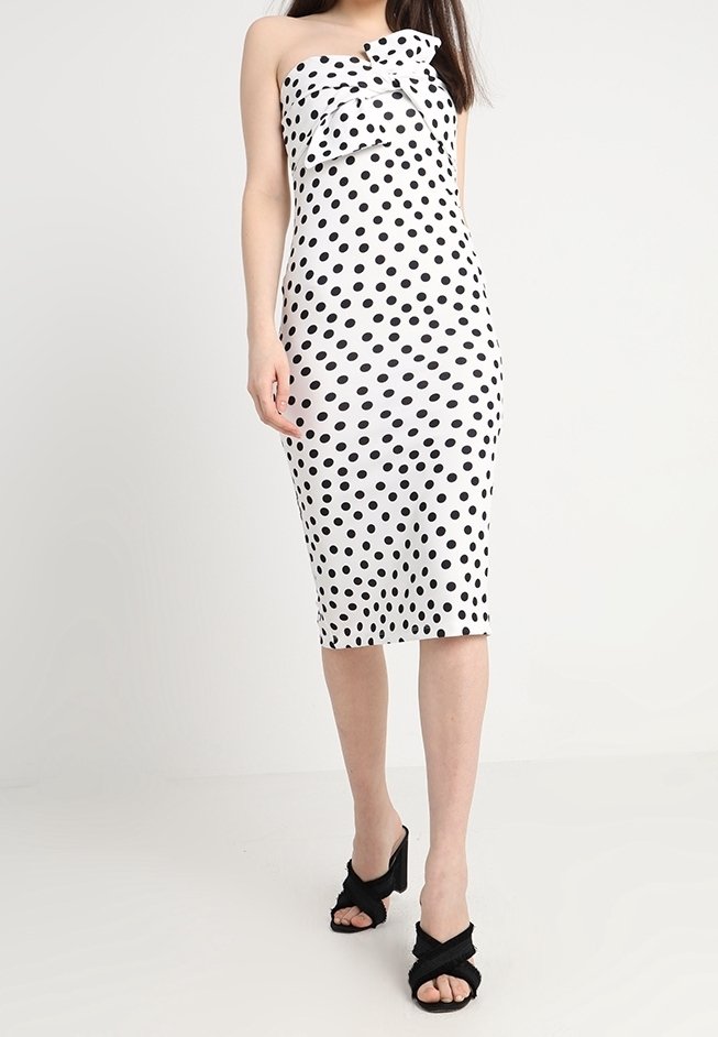 Femme portant une robe midi blanche bustier à pois noirs avec un détail nœud, associée à des talons ouverts noirs.