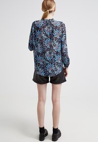 Blus med långa ärmar i svart, blått och lila blommönster, tillsammans med svarta blanka shorts, sedd bakifrån.