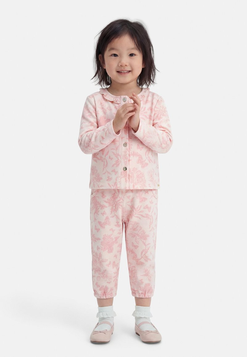 Giovane ragazza in piedi, indossa una maglia a maniche lunghe a fiori rosa e pantaloni coordinati con calzini bianchi alla caviglia e ballerine rosa, sorridente.