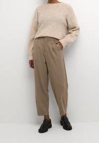 Persoon staat met één hand in de zak, draagt een beige geribbelde trui, lichtbruine hoge taillebroek en zwarte grove loafers tegen een witte muur.