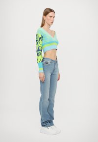 Kratka bluza s V-izrezom v svetlo modri in zeleni barvi, s črtastimi in vzorčenimi rokavi. Svetlo modri jeans in bela športna obutev dopolnjujeta videz.