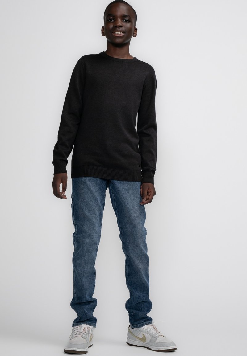 Zwarte gebreide trui met lange mouwen, gecombineerd met lichtblauwe jeans. Draagt witte sneakers met beige accenten. Eenvoudige, casual outfit.