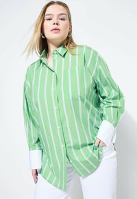 Maglietta a maniche lunghe a righe verdi e bianche con colletto, dotata di taschino frontale e polsini bianchi, indossata con pantaloni bianchi. Texture liscia.