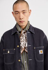 Dark denim jacket with gold buttons, paired with a striped green shirt and a light floral scarf. Features branded pocket detail.