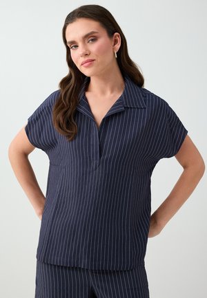 Femme aux longs cheveux bruns portant un chemisier bleu marine à manches courtes et un pantalon assorti à rayures blanches verticales, debout les mains sur les hanches.