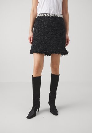 Femme portant une mini-jupe noire texturée avec une ceinture ornée de perles, associée à des bottes noires à talons montant jusqu'aux genoux et un top blanc sans manches.