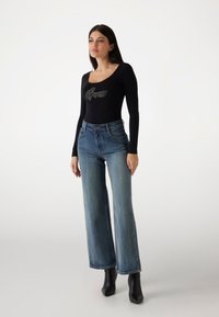 Black long-sleeve bodysuit with a scoop neckline and gray logo, paired with high-waisted blue wide-leg jeans and black ankle boots.