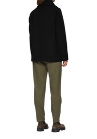 Schwarzer Mantel mit gerader Silhouette und weitem Kragen, kombiniert mit olivgrünen Hosen. Die Schuhe bestehen aus braunen Lederstiefeln.