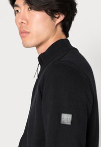 Zwarte gebreide trui met een hoge kraag en ritssluiting, voorzien van een leren logo patch op de linkermouw. Textuurdetails van de stof zijn zichtbaar.