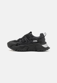 Černé tenisky s robustním, texturovaným designem. Mají síťovinové panely, silné gumové podrážky a stříbrný akcent na patě s tkaničkami pro upevnění.