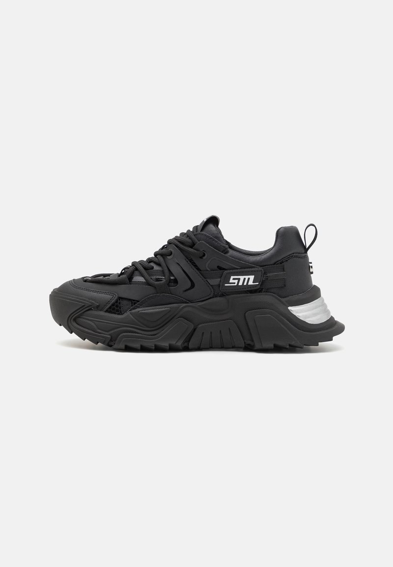 Černé tenisky s robustním, texturovaným designem. Mají síťovinové panely, silné gumové podrážky a stříbrný akcent na patě s tkaničkami pro upevnění.