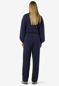 Tumši zila sporta uzvalks ar brīvu piegriezumu, bumbierveida piedurknēm un elastīgu jostasvietu; materiāls izskatās mīksts un ērts.