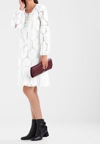 Žena v bílé krajkové šaty, držící tmavě červenou kabelku, oblečená v černých kotníkových botách s květinovými aplikacemi.