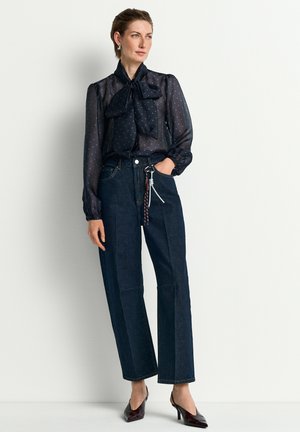 Woman wearing sheer navy blouse with bow tie, high-waisted dark wide-leg jeans, and dark pointed heels, standing against a plain light wall.
