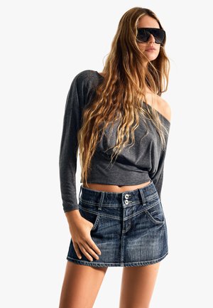 Woman with long wavy hair wearing oversized dark sunglasses, gray off-shoulder long-sleeve top, and a blue denim mini skirt stands with one hand in pocket.