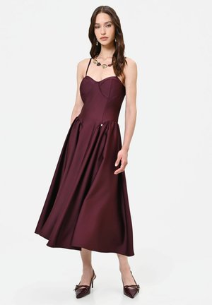 Donna che indossa un abito midi viola scuro con spalline sottili, abbinato a tacchi alti appuntiti coordinati e una collana con grandi pietre.