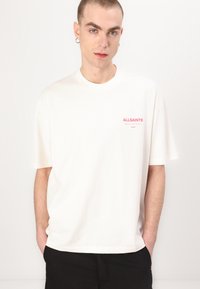 T-shirt en coton blanc avec des manches courtes et un col rond. Présente un petit logo "ALLSAINTS" rose sur la poitrine. Coupe simple et décontractée.