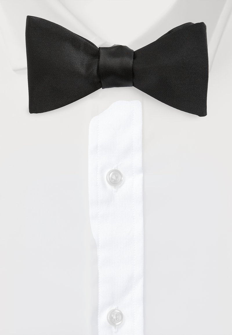 Schwarze Satin-Fliege mit einem zentralen Knoten, die über einem weißen Hemd mit klaren Knöpfen und einem spitzen Kragen sitzt.