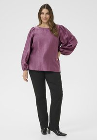 Donna che indossa una blusa lunga viola lucida con maniche a sbuffo, pantaloni neri dritti e stivali neri con tacco, in posa con una mano sulla vita.