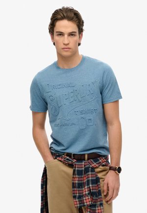 Camiseta de algodón azul con un estampado gráfico texturizado que dice "Original Superdry", combinada con pantalones beiges y una camisa de cuadros atada a la cintura.