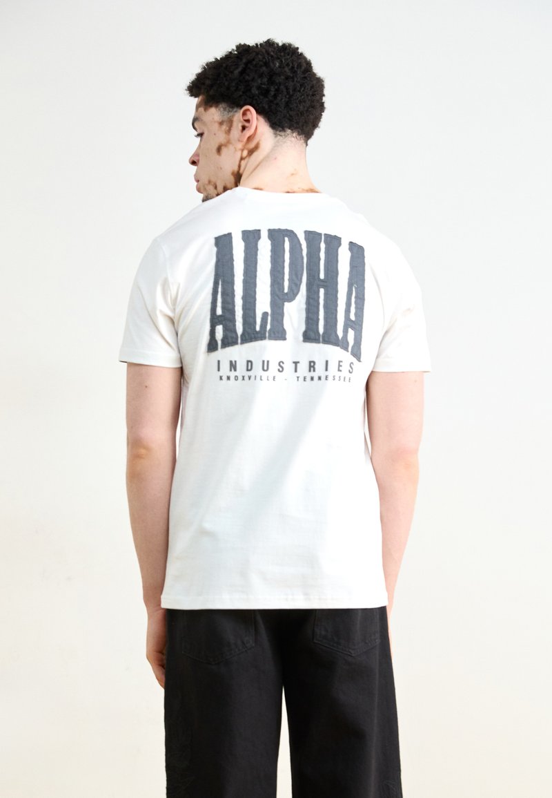 Hvit bomull t-skjorte med en stor grå "ALPHA" grafikk og "INDUSTRIES" tekst på baksiden. Kortermet og design med rund hals.