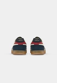 Une paire de baskets basses avec des panneaux beiges, navy et rouges, des semelles en caoutchouc, et "1973" imprimé sur les languettes du talon.