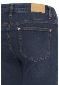 Tumši zili džinsa bikses no muguras ar izšūtu kabatu, vara kniedēm, tumši brūnu ādas zīmola etiķeti uz jostasvietas un redzamām jostas cilpām.