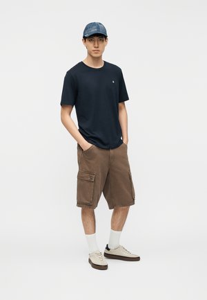 Ung mand iført en marineblå t-shirt, brune cargoshorts, hvide sokker, beige sneakers og en blå denimkasket, stående med hænderne i lommerne.