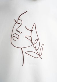 La stoffa bianca presenta un disegno minimalista a linee di un volto e foglie, ricamato con filo marrone. Ricamo che aggiunge texture e dettagli.