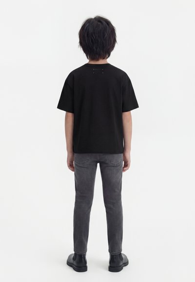 Παιδί με κοντά μαύρα μαλλιά, φοράει μαύρο μπλουζάκι, γκρι τζιν και μαύρες μπότες, στέκεται γυρισμένο με την πλάτη σε λευκό φόντο.