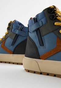 Hoge sneakers met een blauwe bovenkant, grijze en bruine accenten, gele veters, een gestructureerde zool en een zijrits sluiting.