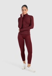 Bordeauxkleurig sportjack met rits en aansluitende lange mouwen, gecombineerd met bijpassende legging; gladde textuur, minimalistisch ontwerp, atletische pasvorm.