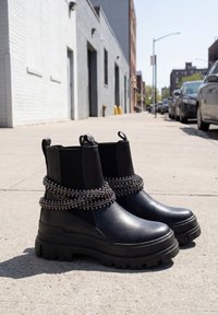 Paire de bottines Chelsea noires épaisses avec des ornements en chaîne argentée posées sur un trottoir urbain avec des bâtiments et des voitures garées en arrière-plan.