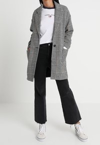 Kappa med hundtandsmönster i svart och vitt, med lång design, uppvikta slag, två knappar och sidofickor, kombinerad med svarta utsvängda byxor och vita sneakers.