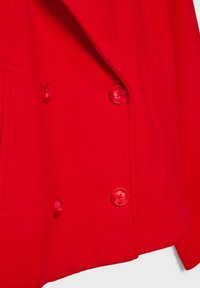 Manteau en laine rouge à double boutonnage avec quatre grands boutons et deux poches verticales fendues, présenté coupé à la taille.