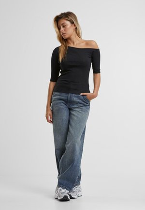 Un top negru off-shoulder cu mâneci trei sferturi, asortat cu blugi largi de culoare albastră. Modelul stă cu o mână în buzunar, purtând adidași gri.