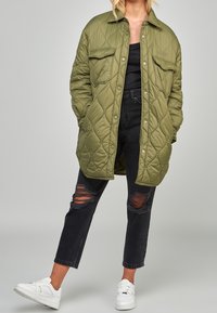 Chaqueta acolchada verde oliva con cuello tipo solapa, botones a presión y dos bolsillos frontales; llevada sobre una camiseta negra y jeans negros desgastados, combinada con zapatillas blancas.