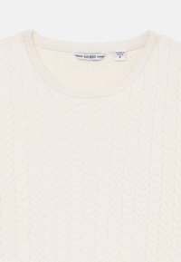 Kremowy sweter z wzorem warkoczy, okrągłym dekoltem i prążkowanym wykończeniem. Metka z napisem "GUESS", wskazująca markę. Wykonany z miękkiego materiału.