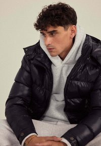 Veste matelassée noire à finition brillante, avec capuche et fermeture éclair frontale, portée sur un sweat à capuche gris clair, associée à un pantalon de survêtement gris.