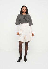 Cardigã cinzento curto com fecho de botão, blusa cremosa de mangas longas, calções plissados bege, collants pretos e tacões pretos. Pequena mala branca.