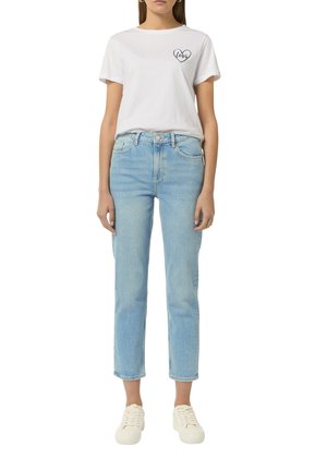 Kvinde iført en hvid t-shirt med et hjerte og teksten "kærlighed" på brystet, lyseblå højttaljede jeans og hvide sneakers, stående oprejst.